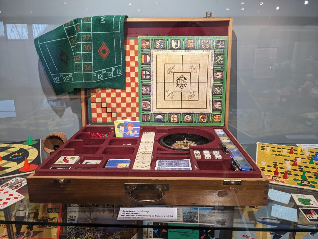 in einer Vitrine steht ein Spielekoffer, verschieden Spielbretter, Spielsteine, Würfel und Spielkarten