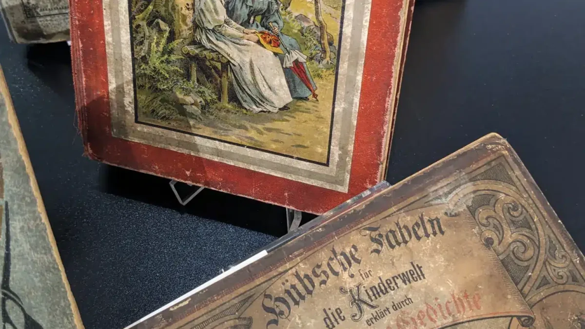 Zwei alte Bücher mit bunten, illustrierten Buchcovern stehen auf einem Tisch in einem Ausstellungsraum.