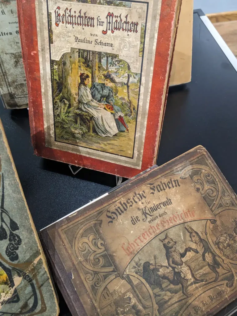 Zwei alte Bücher mit bunten, illustrierten Buchcovern stehen auf einem Tisch in einem Ausstellungsraum.