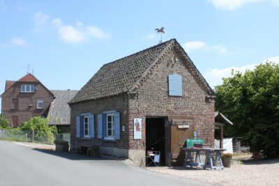 Ein kleines, gemauertes Haus mit einem Schopf und einem Pferd auf der Wetterfahne, umgeben von einer ländlichen Kulisse.