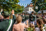 Festliche Umzugsszene mit Menschen in Trachten, die eine Fahne halten und zur Parade auf der Dorfstraße winken.