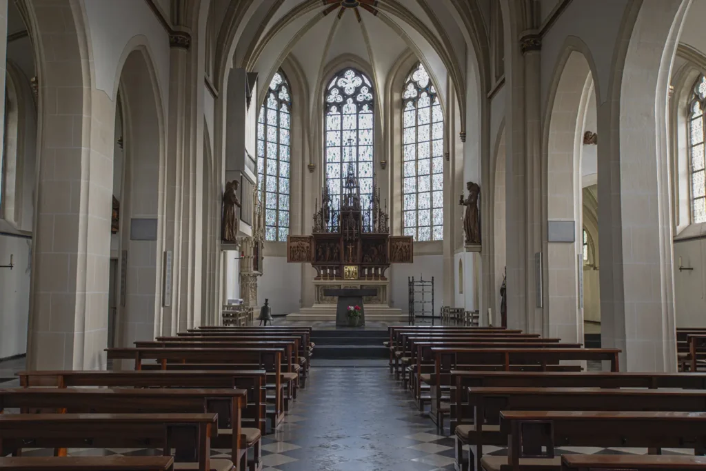 Innenansicht einer Kirche mit hohen Gewölben, bunten Fenstern und Holzbänken für die Gottesdienstbesucher.