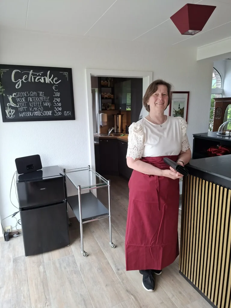 Eine Frau in einem weißen Oberteil und roter Schürze steht lächelnd an einem Empfangstresen in einem gemütlichen Raum.
