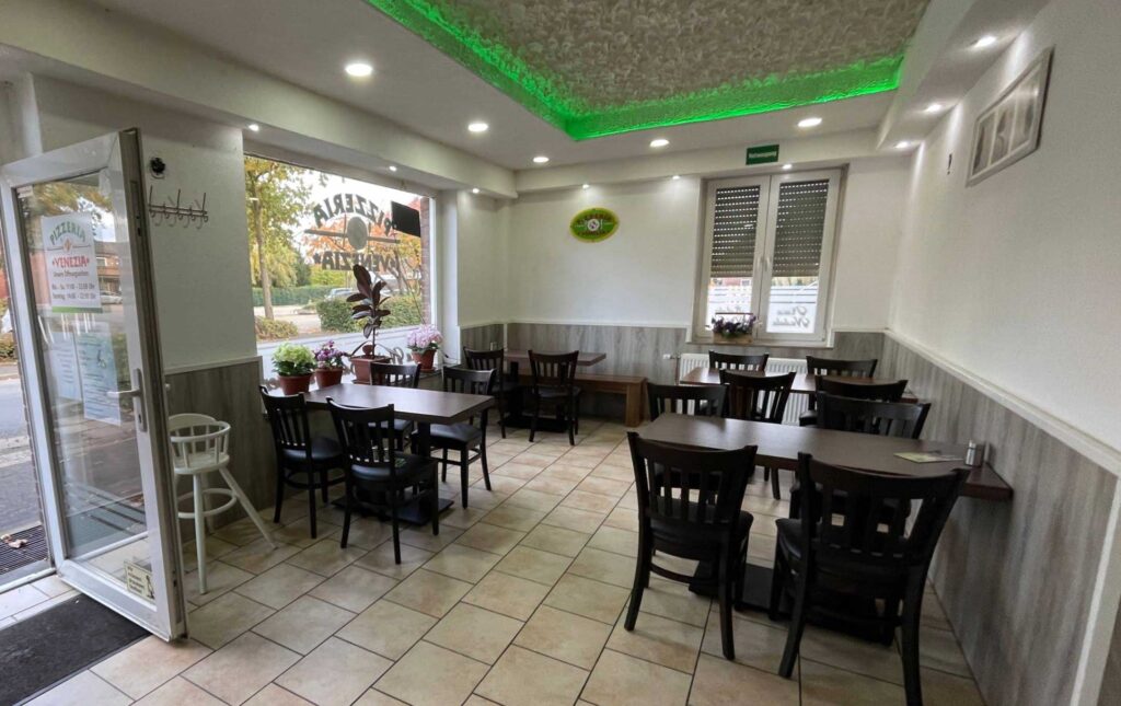 Ein helles Restaurant mit Tischen, Stühlen und Fensterpflanzen, die gemütliche Atmosphäre schafft.