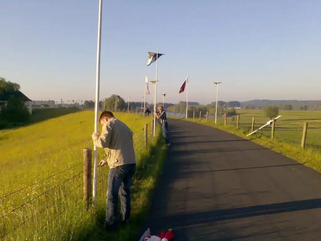 Zwei Personen befestigen Fahnen an Pfählen entlang eines Radwegs in ländlicher Umgebung am frühen Morgen.