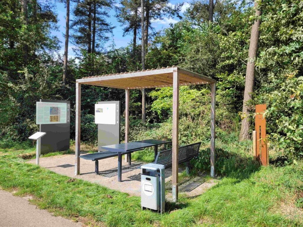 Rastplatz mit überdachten Tisch und Bank, Infotafeln und Abfallbehälter, umgeben von Bäumen und Wiese.